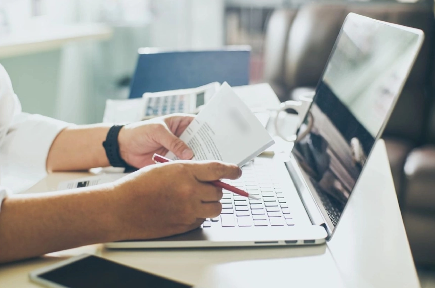 A person holding a paper and typing on a laptop.