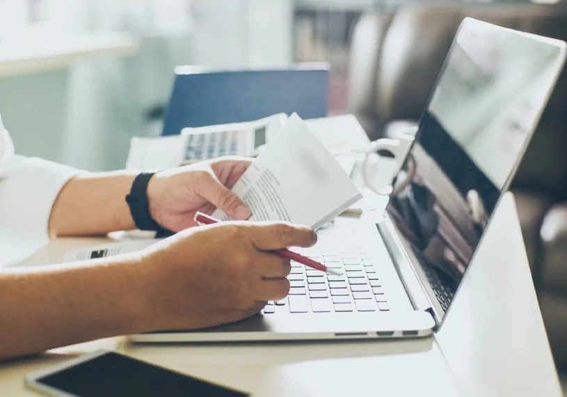 A person holding a paper and typing on a laptop.